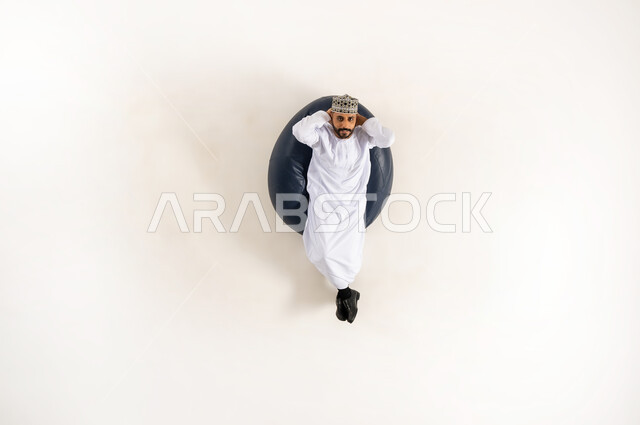 الاسترخاء والتأمل في اوقات الفراغ، متعة قضاء الوقت مع الذات، بورتريه عامودي علوي لرجل عربي خليجي عماني يرتدي الدشداشة والكمه مستلقي على كنبه مريحه ويضع يديه خلف رأسه و ينظر الى الأعلى، ايماءات تدل على الراحة والسرور، خلفية بيضاء