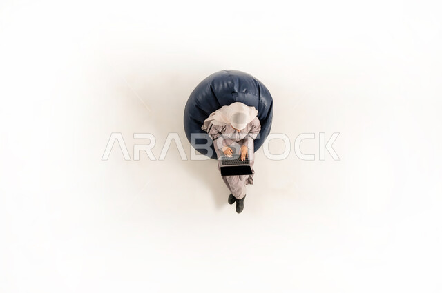 Integration and focus to accomplish tasks remotely, performing practical tasks and duties, vertical upper portrait of an Arab Gulf Omani lady wearing hijab and abaya sitting on a comfortable sofa and working on a laptop, Saudi woman using modern technological devices, white background