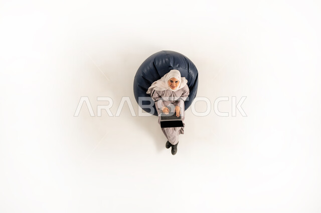 Performing practical tasks and duties, integration and focus to accomplish tasks remotely, vertical upper portrait of an Arab Gulf Omani woman wearing hijab and abaya sitting on a chair and looking up working on a laptop, Saudi woman using modern technological devices, white background