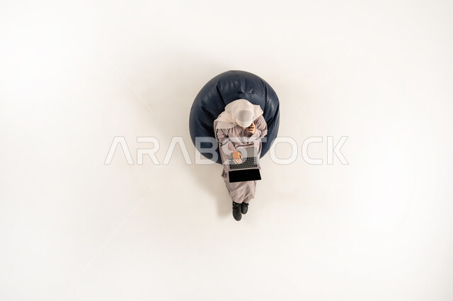 Working on laptop, performing practical tasks and duties, integrating and concentrating to accomplish tasks remotely, vertical upper portrait of an Arab Gulf Omani lady wearing hijab and abaya sitting on a comfortable chair holding a phone in her hand, using modern technological devices, white background