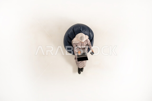 العمل على جهاز الحاسب المحمول، امراة سعودية تؤدي المهام والواجبات العملية، الاندماج والتركيز لإنجاز المهام عن بعد، بورتريه عامودي علوي لسيده عربية خليجية عمانية ترتدي الحجاب والعباءه تجلس على كرسي مريح تحمل الهاتف بيدها، استخدام الأجهزة التكنولوجية الحديثة، خلفيه بيضاء