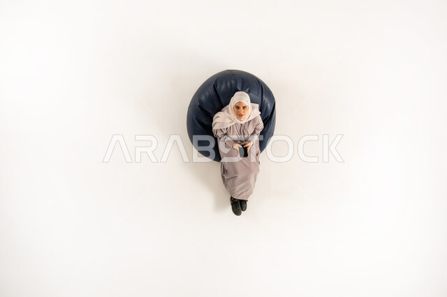Using mobile phone in free time, Saudi woman using modern devices to communicate with family and friends, vertical top portrait of a veiled Gulf Omani Arab woman wearing an abaya sitting on a comfortable sofa looking up holding her mobile device, white background