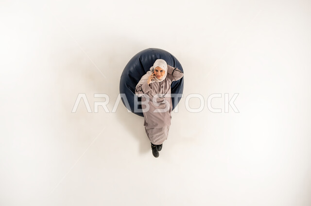 التواصل مع الأهل والأصدقاء، سيدة سعودية تتبادل الأحاديث والأخبار الجيدة، الاتصال الهاتفي عبر الجوال، بورتريه عامودي علوي لشابة عربية خليجية عمانية ترتدي العباءه والحجاب تجلس على كنبه مريحه وتنظر الى الاعلى تقوم بإجراء مكالمة هاتفية عبر الهاتف المحمول، خلفية بيضاء