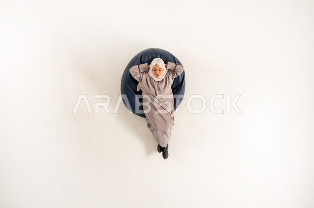 مفهوم الاسترخاء والاستجمام، سيدة سعودية بحاجة إلى الراحة و الهدوء، بورتريه علوي عامودي لأمرأة عربية خليجية عمانية تجلس على كنبة مريحة وتقوم بوضع يديها خلف راسها وتنظر الى الاعلى، خلفية بيضاء