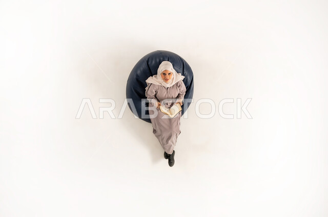 Exploiting time to educate yourself, the importance of reading in Omani society, a vertical portrait of a veiled Omani Gulf Arab woman wearing an abaya and looking up, holding a book in her hand, sitting on a comfortable sofa reading, white background