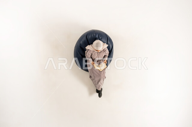 أهمية المطالعة في المجتمع العماني,  امراة سعودية تستغل الوقت في التثقف، بورتريه عامودي علوي لسيدة عربية خليجية عمانية محجبة ترتدي العباءة و تحمل بيدها كتابا تجلس على كنبه مريحه تقوم بالقراءه, خلفية بيضاء