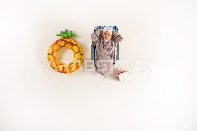 الراحه والاستجمام في العطله الصيفيه، مفهوم الأناقة والاهتمام بالمظهر الخارجي، بورتريه عامودي علوي لسيدة عربية خليجية عمانية ترتدي العباءة والحجاب، امراة سعودية تجلس على كرسي الشاطئ و تنظر الى الاعلى، خلفيه بيضاء