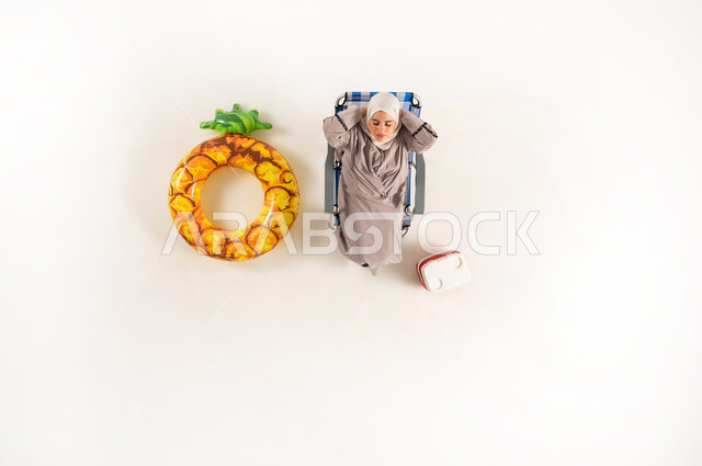 مفهوم الأناقة والاهتمام بالمظهر الخارجي، الراحه والاستجمام في العطله الصيفيه، بورتريه عامودي علوي لسيدة عربية خليجية عمانية ترتدي العباءة والحجاب، امراة سعودية تجلس على كرسي الشاطئ و تنظر الى الاعلى، خلفيه بيضاء