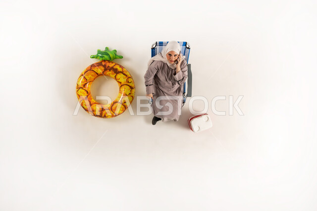 Using mobile phones in free time, using modern devices to communicate with family and friends, the concept of comfort and recreation during summer vacation, vertical upper portrait of a veiled Gulf Arab Omani woman wearing an abaya sitting on a beach chair talking on a mobile phone, white background