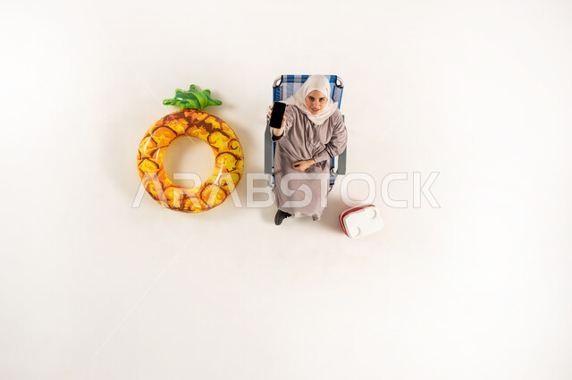 استعمال الاجهزه الحديثه في التواصل مع الاهل والاصدقاء، مفهوم الراحه والاستجمام في العطله الصيفيه، بورتريه عامودي علوي لسيده عربيه خليجيه عمانية محجبه ترتدي العباءه  تجلس على كرسي الشاطئ تقوم بعرض هاتف محمول بشاشة سوادء، استخدام الجوال في اوقات الفراغ، خلفيه بيضاء