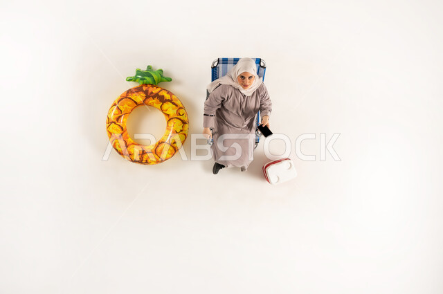 استخدام الجوال في اوقات الفراغ، امراة سعودية تستخدم الاجهزه الحديثه في التواصل مع الاهل والاصدقاء، مفهوم الراحه والاستجمام في العطله الصيفيه، بورتريه عامودي علوي لسيده عربيه خليجيه عمانية محجبه ترتدي العباءه تجلس على كرسي الشاطئ تقوم بتصفح الهاتف المحمول، خلفيه بيضاء