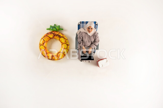 Performing practical tasks and duties, integration and focus to accomplish tasks remotely, vertical top portrait of an Arab Gulf Omani woman wearing hijab and abaya sitting on a chair and looking up working on a laptop, Saudi woman using modern technological devices, white background