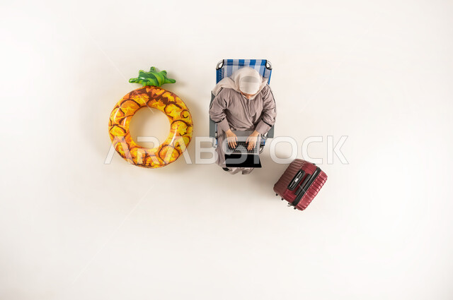 الاندماج والتركيز لإنجاز المهام عن بعد، بورتريه عامودي علوي لسيده عربية خليجية عمانية ترتدي الحجاب والعباءه تجلس على كرسي تقوم بالعمل على جهاز الحاسب المحمول، امراة سعودية تؤدي المهام والواجبات العملية، استخدام الأجهزة التكنولوجية الحديثة، خلفيه بيضاء