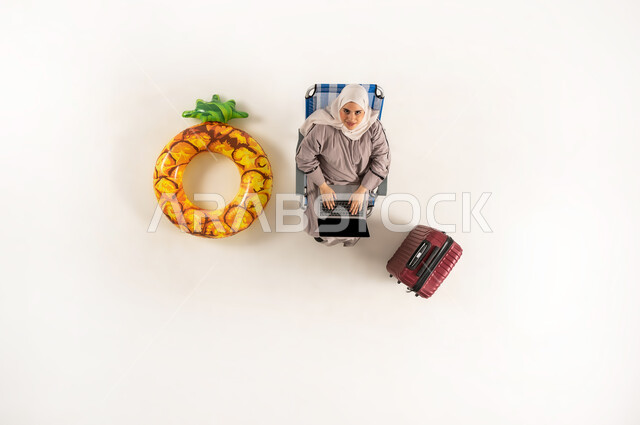 Performing practical tasks and duties, Saudi woman merging and concentrating to accomplish tasks remotely, vertical top portrait of an Arab Gulf Omani lady wearing hijab and abaya sitting on a chair and looking up working on a laptop, using modern technological devices, white background