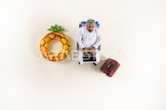 الجلوس و العمل على الحاسوب المحمول، الاستفادة من التكنولوجيا والتقنيات المتطورة، بورتريه عامودي علوي لرجل عربي خليجي عماني يرتدي الدشداشة والكمه وينظر الى الاعلى، قضاء الوقت مع الذات، انجاز المهام عن بعد، خلفية بيضاء