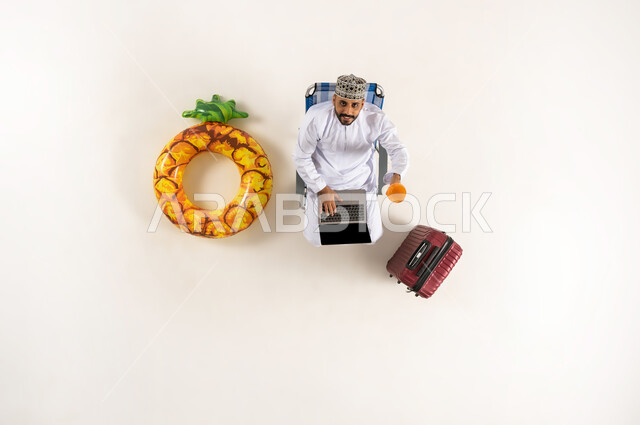 الجلوس و العمل على الحاسوب المحمول، الاستفادة من التكنولوجيا والتقنيات المتطورة، بورتريه عامودي علوي لرجل عربي خليجي عماني يرتدي الدشداشة والكمه يشرب العصير المنعش وينظر الى الاعلى، قضاء الوقت مع الذات، انجاز المهام عن بعد، خلفية بيضاء