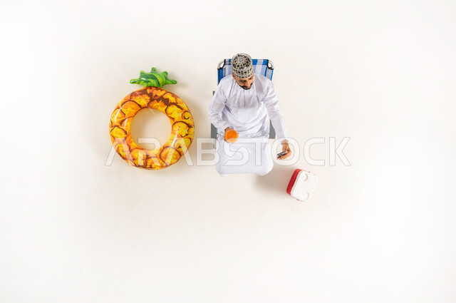 مفهوم الراحه والاستجمام في العطله الصيفيه، قضاء انشطة ترفيهية ممتعة مع الذات, الاستمتاع بالمشروبات المنعشة,  بورتريه علوي عامودي لرجل عربي خليجي عماني يرتدي الدشداشة والكمه  يجلس على كرسي الشاطئ، خلفية بيضاء