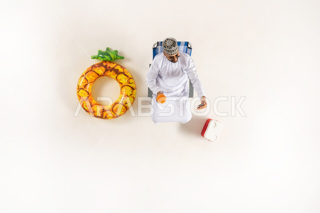 قضاء انشطة ترفيهية ممتعة مع الذات, مفهوم الراحه والاستجمام في العطله الصيفيه، الاستمتاع بالمشروبات المنعشة,  بورتريه علوي عامودي لرجل عربي خليجي عماني يرتدي الدشداشة والكمه  يجلس على كرسي الشاطئ، خلفية بيضاء