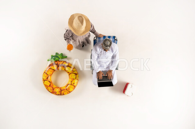 الاستمتاع بالمشروبات الصيفية المنعشة, قضاء انشطة ترفيهية ممتعة برفقة الزوجة,  بورتريه علوي عامودي لزوج عربي خليجي عماني يرتدي الدشداشة والكمه ويضع النظارات الشمسية يعمل على الحاسب المحمول,  خلفية بيضاء