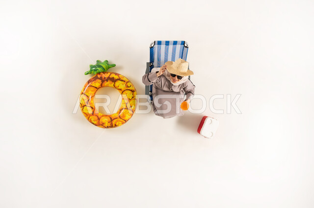 استخدام الجوال في اوقات الفراغ، امراة سعودية تستخدم الاجهزه الحديثه في التواصل مع الاهل والاصدقاء، مفهوم الراحه والاستجمام في العطله الصيفيه، بورتريه عامودي علوي لسيده عربيه خليجيه عمانية محجبه ترتدي العباءه تجلس على كرسي الشاطئ تقوم بتصفح الهاتف المحمول، خلفيه بيضاء