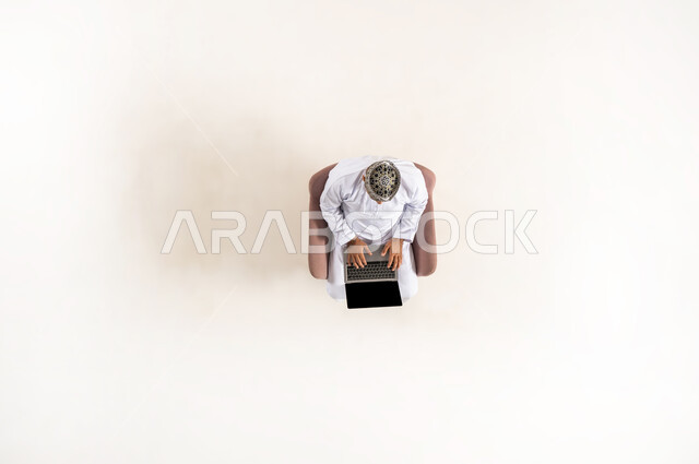 الاستفادة من التكنولوجيا والتقنيات المتطورة، بورتريه عامودي علوي لرجل عربي خليجي عماني يرتدي الدشداشة والكمه، الجلوس و العمل على الحاسوب المحمول، قضاء الوقت مع الذات، انجاز المهام عن بعد، خلفية بيضاء