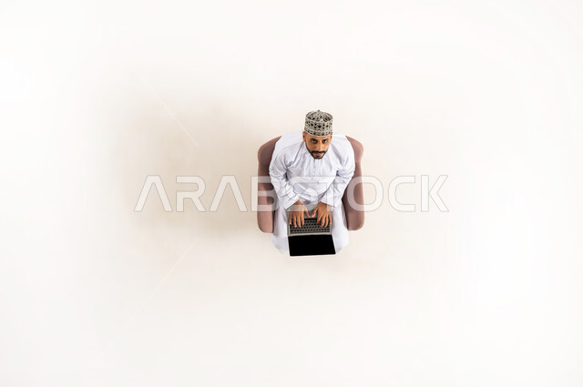 Sitting and working on laptop, taking advantage of technology and advanced techniques, vertical upper portrait of an Arab Gulf Omani man wearing a dishdasha and a turban and looking up, spending time with oneself, completing tasks remotely, white background
