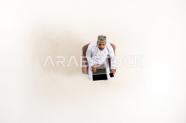 Sitting and working on laptop, taking advantage of technology and advanced techniques, vertical upper portrait of an Arab Gulf Omani man wearing a dishdasha and a turban and looking up, spending time with oneself, completing tasks remotely, white background