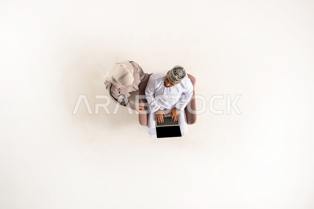 الاستفادة من التكنولوجيا والتقنيات المتطورة، انجاز المهام عن بعد، بورتريه عامودي علوي لزوج عربي خليجي عماني يرتدي الدشداشة والكمه برفقة زوجته، الجلوس و العمل على الحاسوب المحمول، قضاء الوقت مع الزوجة، خلفية بيضاء