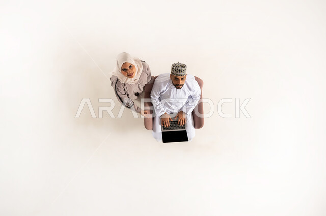 Completing tasks remotely, taking advantage of advanced technology and techniques, vertical upper portrait of an Arab Gulf Omani couple wearing a dishdasha and a turban, looking up with his wife, sitting and working on a laptop, spending time with his wife, white background