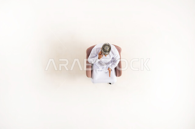 Using modern technology and techniques, e-marketing and displaying mobile services and applications, vertical upper portrait of an Arab Gulf Omani man wearing a dishdasha and a turban sitting on a comfortable sofa and talking on a mobile phone, white background