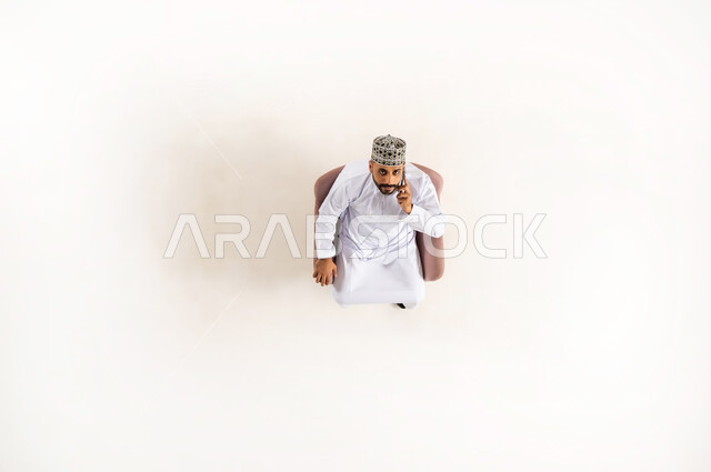 اجراء مكالمة عبر الهاتف المحمول، إستخدام التكنولوجيا والتقنية الحديثة، التواصل مع الاهل والاصدقاء، بورتريه عامودي علوي لرجل عربي خليجي عماني يرتدي الدشداشة والكمه يجلس على كنبه مريحه وينظر الى الاعلى، خلفية بيضاء
