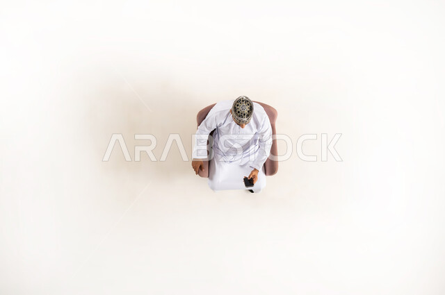 التسويق الإلكتروني وعرض خدمات وتطبيقات الجوال، إستخدام التكنولوجيا والتقنية الحديثة، بورتريه عامودي علوي لرجل عربي خليجي عماني يرتدي الدشداشة والكمه يجلس على كنبه مريحه ويتصفح الهاتف المحمول، خلفية بيضاء