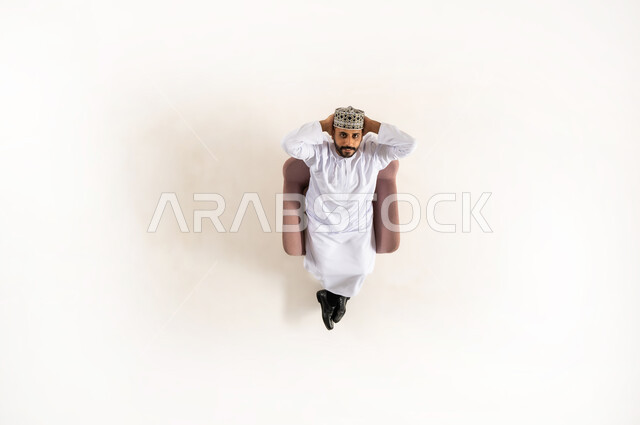 الاسترخاء والتأمل في اوقات الفراغ، متعة قضاء الوقت مع الذات، بورتريه عامودي علوي لرجل عربي خليجي عماني يرتدي الدشداشة والكمه مستلقي على كنبه مريحه ويضع يديه خلف رأسه و ينظر الى الأعلى، ايماءات تدل على الراحة والسرور، خلفية بيضاء