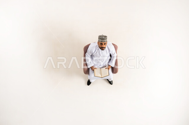 استغلال الوقت في التثقف، مفهوم التمعن والتركيز، أهمية المطالعة في المجتمع العماني,  قضاء الوقت بالقراءة والتعلم والثقافة ، بورتريه عامودي علوي لرجل عربي عماني خليجي يرتدي الدشداشة والكمه يجلس على كرسي مريح  ينظر الى الاعلى ويحمل بيديه كتاب، خلفية بيضاء