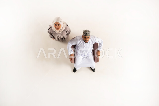 قضاء وقت ممتع في المنزل مع العائلة، بورتريه علوي عامودي لرجل عربي خليجي عماني يرتدي الدشداشة ويضع الكمة يجلس على كنبه وينظر الى الكاميرا هو وزوجته، الاستمتاع بالأنشطة العائلية الترفيهيه في عطلة نهاية الاسبوع, أجواء مليئة بالحب، خلفية بيضاء