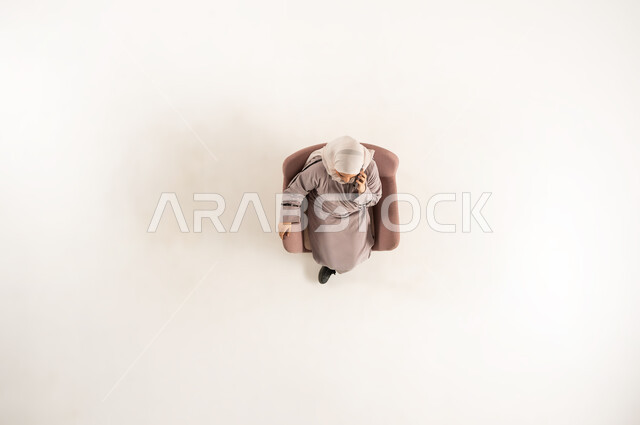تبادل الأحاديث والأخبار الجيدة، التواصل مع الأهل والأصدقاء، الاتصال الهاتفي عبر الجوال، بورتريه عامودي علوي لسيدة عربية خليجية عمانية ترتدي العباءه والحجاب تجلس على كنبه مريحه من الجلد، القيام بإجراء مكالمة هاتفية عبر الهاتف المحمول، خلفية بيضاء