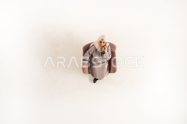 التواصل مع الأهل والأصدقاء، امراة سعودية تتبادل الأحاديث والأخبار الجيدة، الاتصال الهاتفي عبر الجوال، بورتريه عامودي علوي لسيدة عربية خليجية عمانية ترتدي العباءه والحجاب تجلس على كنبه مريحه من الجلد وتنظر الى الاعلى، القيام بإجراء مكالمة هاتفية عبر الهاتف المحمول، خلفية بيضاء