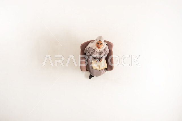 استغلال الوقت في التثقف، أهمية المطالعة في المجتمع العماني, بورتريه عامودي علوي لسيدة عربية خليجية عمانية محجبة ترتدي العباءة وتنظر الى الاعلى تحمل بيدها كتابا، امراة سعودية تجلس على كنبه مريحه تقوم بالقراءه, خلفية بيضاء