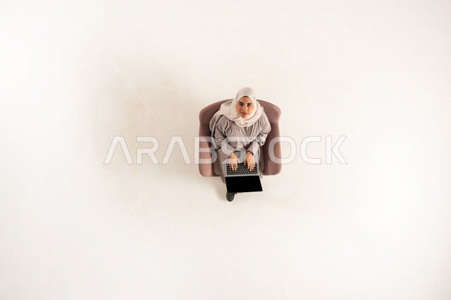 امراة تقوم بأداء الواجبات العملية، الاندماج والتركيز لإنجاز المهام عن بعد، بورتريه عامودي علوي لسيده عربية خليجية عمانية ترتدي الحجاب والعباءه وتنظر الى الاعلى تجلس على كرسي مريح تبدأ بالعمل على جهاز الحاسب المحمول، استخدام الأجهزة التكنولوجية الحديثة، خلفيه بيضاء