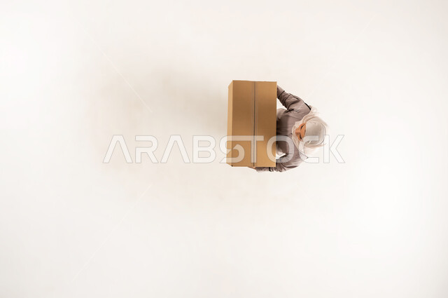 عملية توصيل الطلبات للزبائن بالوقت المحدد وبشكل سليم، سيدة سعودية تتسوق وتطلب المنتجات عبر الانترنت، خدمة الشحن والتوصيل ذات الجودة العالية، بورتريه علوي عامودي لامراة عربية خليجية عماني محجبة ترتدي العباءة تحمل بيدها صندوق كرتوني، الجسم بالطول الكامل، خلفية بيضاء