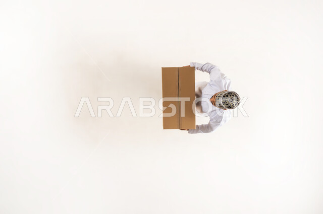 عملية توصيل الطلبات للزبائن بالوقت المحدد وبشكل سليم، خدمة الشحن والتوصيل ذات الجودة العالية، التسوق وطلب المنتجات عبر الانترنت، بورتريه علوي عامودي لرجل عربي خليجي عماني يرتدي الدشداشة والكمه يحمل بيده صندوق كرتوني، الجسم بالطول الكامل، خلفية بيضاء