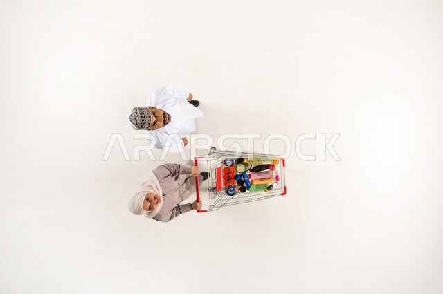 الاستفادة من الخصم و التخفيض على المنتجات، البحث عن المستلزمات المنزلية التموينيه من المركز التجاري العماني, بورتريه عامودي علوي لزوجان عربيان خليجيان عمانيان يقومان بجر عربة التسوق والنظر الى الاعلى، الشراء من السوبر ماركت, خلفيه بيضاء