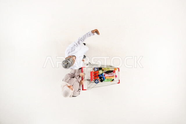 التقاط صورة سيلفي تذكارية عبر الهاتف المحمول، البحث عن المستلزمات المنزلية التموينيه من المركز التجاري العماني, الشراء من السوبر ماركت، الاستفادة من الخصم و التخفيض على المنتجات، بورتريه عامودي علوي لزوجان عربيان خليجيان عمانيان يقومان بجر عربة التسوق, خلفيه بيضاء