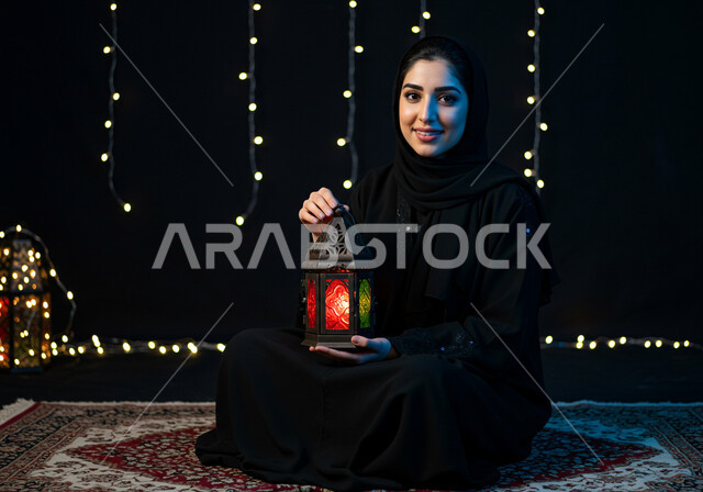 Preparing and getting ready to welcome the holy month of Ramadan, a smiling Saudi Arabian Gulf woman wearing a hijab and abaya holding a decorated illuminated metal lantern sitting on a carpet, holiday and occasion decorations and lights, celebrating the arrival of the month of goodness