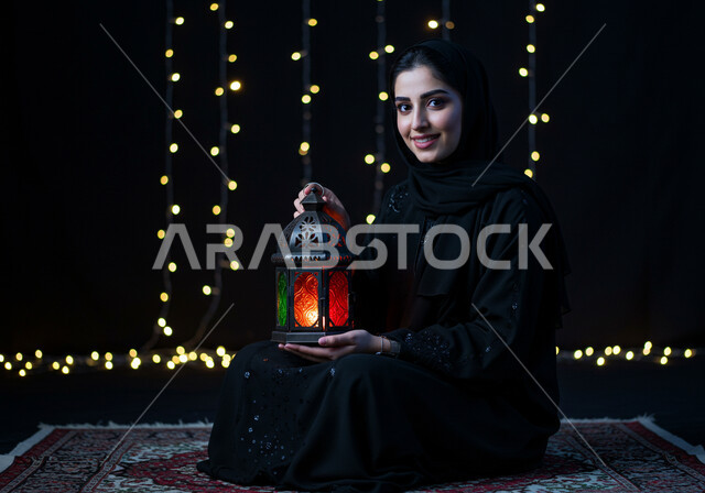  الاستعداد والتجهز لاستقبال شهر رمضان المبارك، رجل عربية خليجية سعودية مبتسمة ترتدي الحجاب والعباءة تمسك فانوس معدني مزخرف مضيء تجلس على السجادة، زينة وأنوار الأعياد والمناسبات، الاحتفال بقدوم شهر الخير