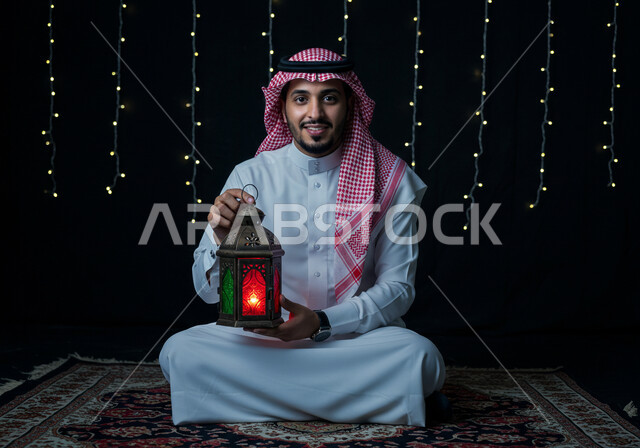 Holiday and occasion decorations and lights, celebrating the arrival of the month of goodness, preparing and getting ready to receive the holy month of Ramadan, a smiling Saudi Arabian Gulf man wearing a traditional shemagh and thobe holding a decorated illuminated metal lantern sitting on the carpet