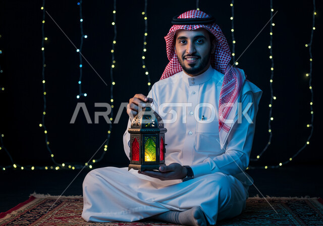  زينة وأنوار الأعياد والمناسبات، الاحتفال بقدوم شهر الخير، الاستعداد والتجهز لاستقبال شهر رمضان المبارك، رجل عربي خليجي سعودي مبتسم يرتدي الشماغ والثوب التقليدي يمسك فانوس معدني مزخرف مضيء يجلس على السجادة