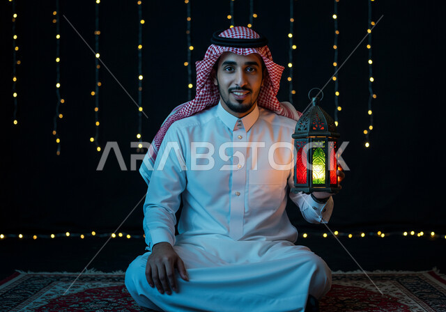 الاحتفال بقدوم شهر الخير، الاستعداد والتجهز لاستقبال شهر رمضان المبارك، رجل عربي خليجي سعودي مبتسم يرتدي الشماغ والثوب التقليدي يمسك فانوس معدني مزخرف مضيء يجلس على السجادة، زينة وأنوار الأعياد والمناسبات 