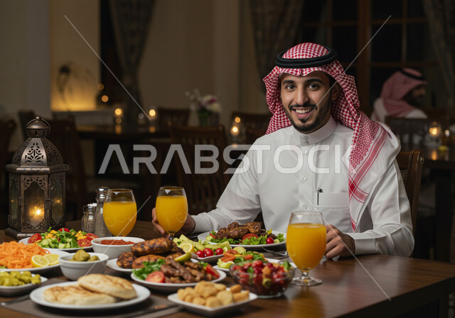 A Ramadan table full of delicious food, appetizers and cold drinks, looking at the camera with expressions of joy and happiness at the arrival of the holy month of Ramadan, a close-up photo of a smiling Saudi Arabian Gulf man wearing a traditional shemagh and thobe sitting around the iftar table at sunset
