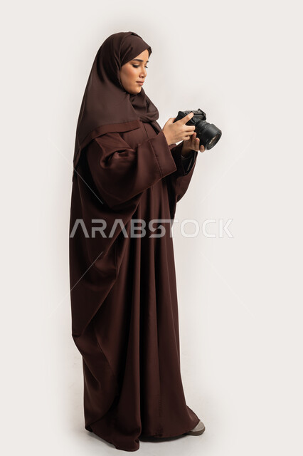 احتراف التصوير الفوتوغرافي باستخدام كاميرات رقمية عالية الدقة, بورتريه لشابة عربية خليجية اماراتية ترتدي الحجاب والعباءة تقوم بالتقاط صورة تذكارية عبر كاميرا فوتوغرافية, مصورة سعودية تمارس هواية التصوير، مهنة ووظيفة نسائية, الجسم بالطول الكامل، خلفية بيضاء